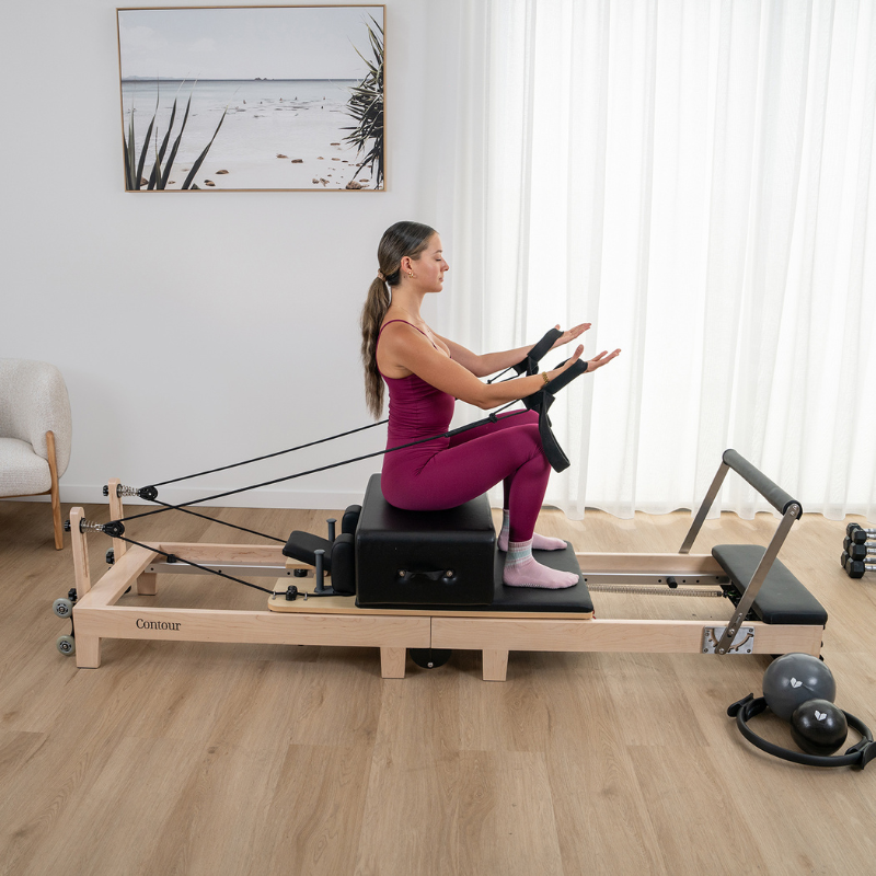 A woman on a Pilates sitting box working out on the Lifespan Fitness Contour Folding Wooden Pilates Reformer Machine