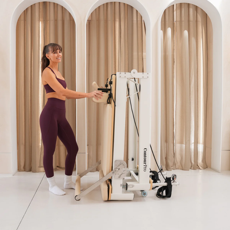 Woman standing by the Lifespan Fitness Contour Pro Aluminium Folding Pilates Reformer Machine in Beige in folded upright position inside a studio room