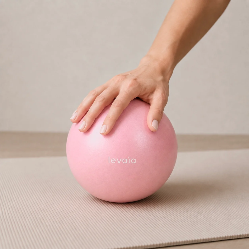 Hand resting on a levaïa luna Soft Pilates Ball in Blush on a beige exercise mat.