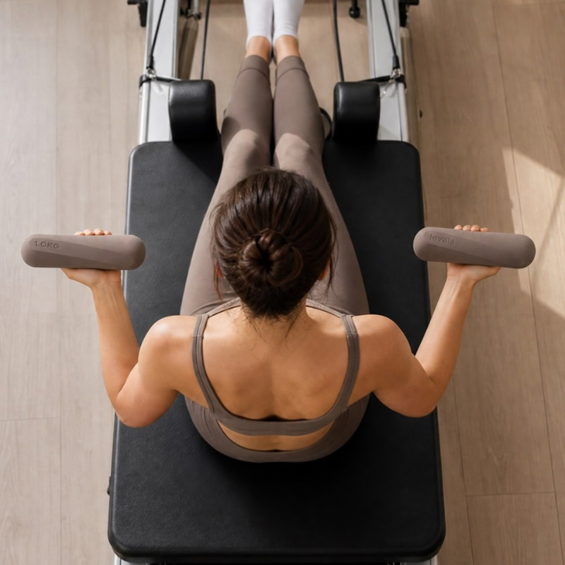 Woman exercising on a Pilates reformer while holding levaïa lifts - Handheld Pilates Weights Pair in Taupe