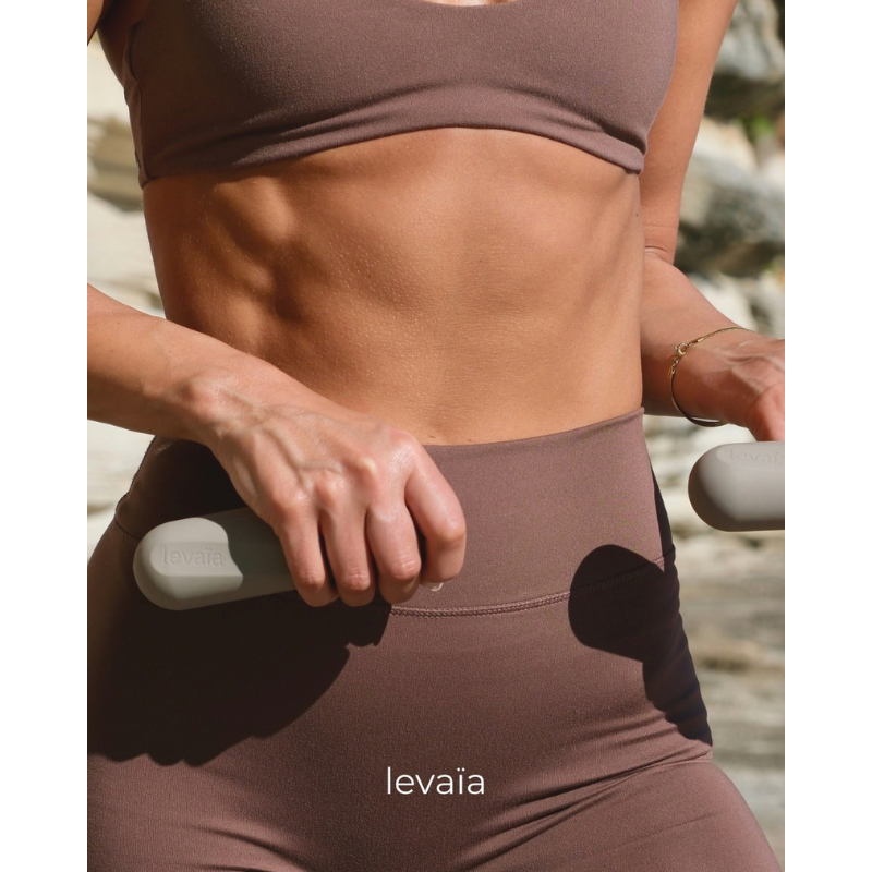 Woman wearing Pilates outfit and holding levaïa lifts - Handheld Pilates Weights Pair in Taupe at the beach.