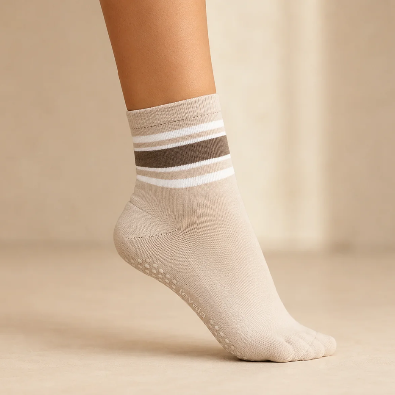 Foot wearing a levaïa Pilates Grip Sock in Taupe, with taupe and white ankle stripes and grip dots visible beneath the raised heel.