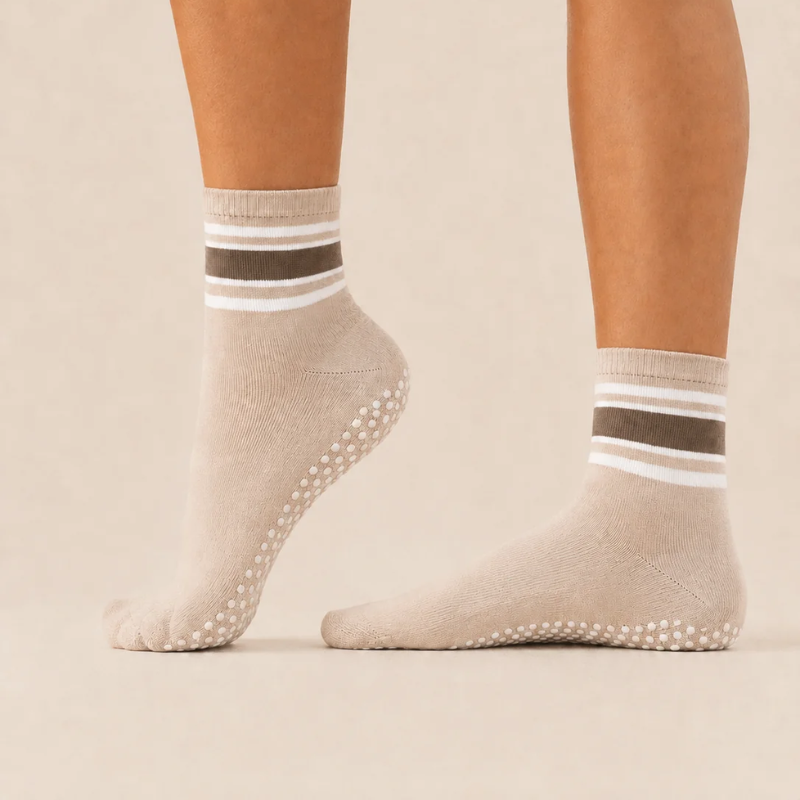 Side view of feet wearing levaïa Pilates Grip Socks in Taupe, with taupe and white ankle stripes and one heel raised to show grip dots.