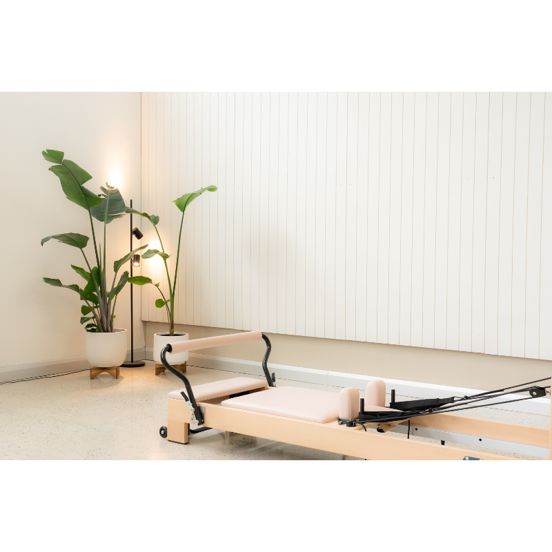 Partial view of the Reeplex Luna Beechwood Pilates Reformer Machine and its carriage inside a studio by plants and lamps.