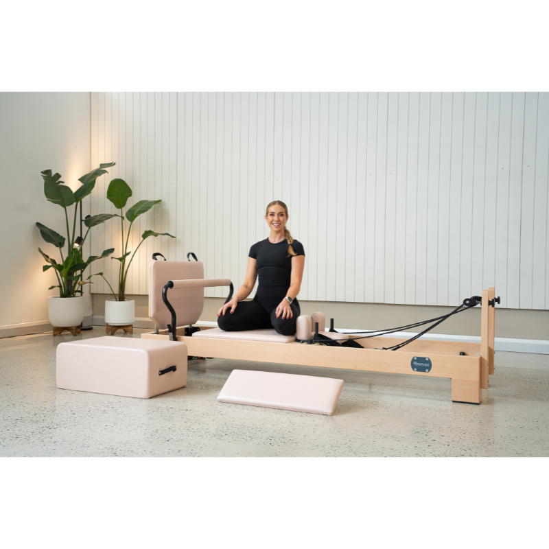Woman sitting on the Reeplex Luna Beechwood Pilates Reformer Machine in a modern studio with plants decor and all reformer accessories laid out in front.