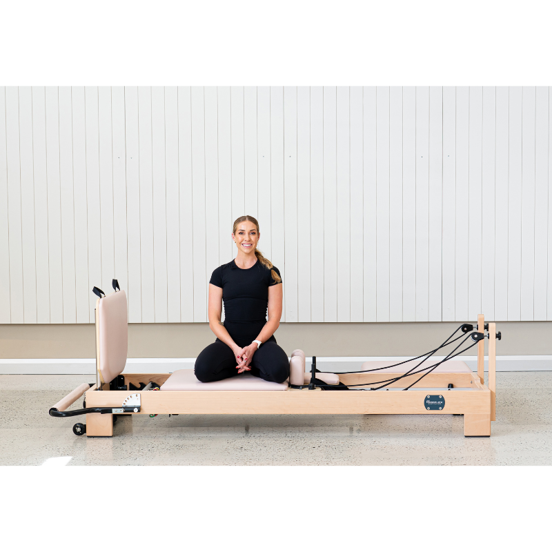Woman sitting on the Reeplex Luna Beechwood Pilates Reformer Machine inside a minimalist studio.